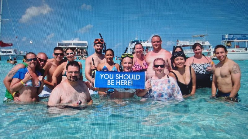 Group of happy Tours Cayman guests standing in turquoise water at Stingray City holding a You Should Be Here sign, Grand Cayman